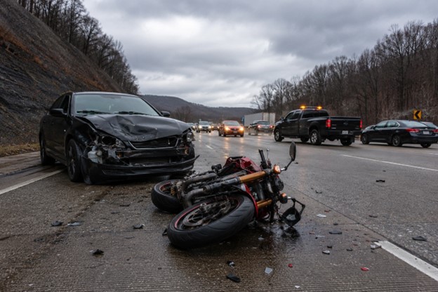 A wrecked red and black motorcycle lies on its side on a damp Pennsylvania highway next to a black sedan with severe front-end damage, with scattered debris on the asphalt and traffic moving past in the background under an overcast sky.