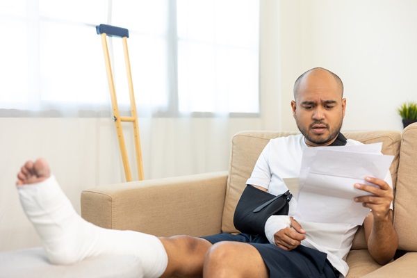A man with a cast on his leg and his arm in a sling sits on a sofa, looking concerned while reviewing medical paperwork. A crutch leans against the wall in the background.