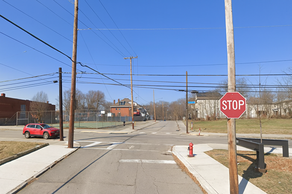 A clear blue sky over a Pittsburgh intersection with a red SUV on the left, a stop sign in the foreground, and a fenced basketball court beside a brick building.
