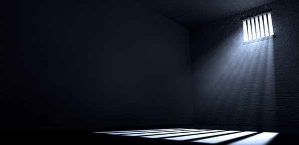 A dimly lit, empty prison cell with dark stone walls and a single small, barred window near the ceiling that allows a dramatic beam of light to stream across the floor.