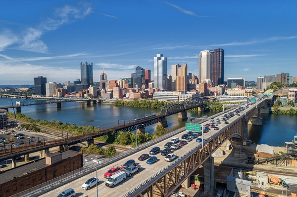 An aerial shot of heavy traffic on a bridge and highway heading toward the downtown Pittsburgh skyline across a river on a sunny day.